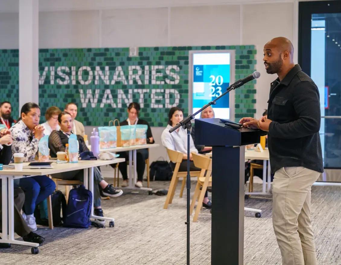 Person stands at a podium addressing a seated audience, with a sign in the background that reads: Visonaries Wanted