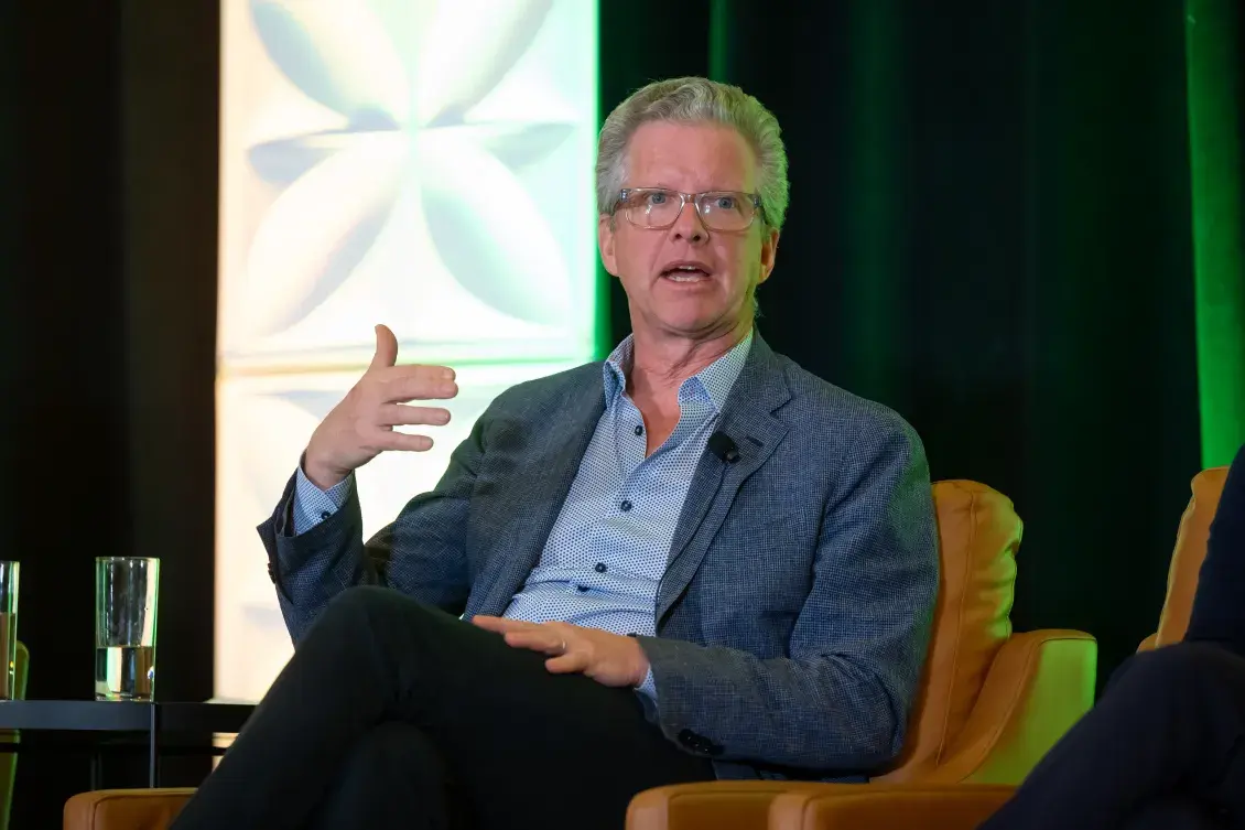 Person in blue shirt and jacket speaks, gesturing with their hand
