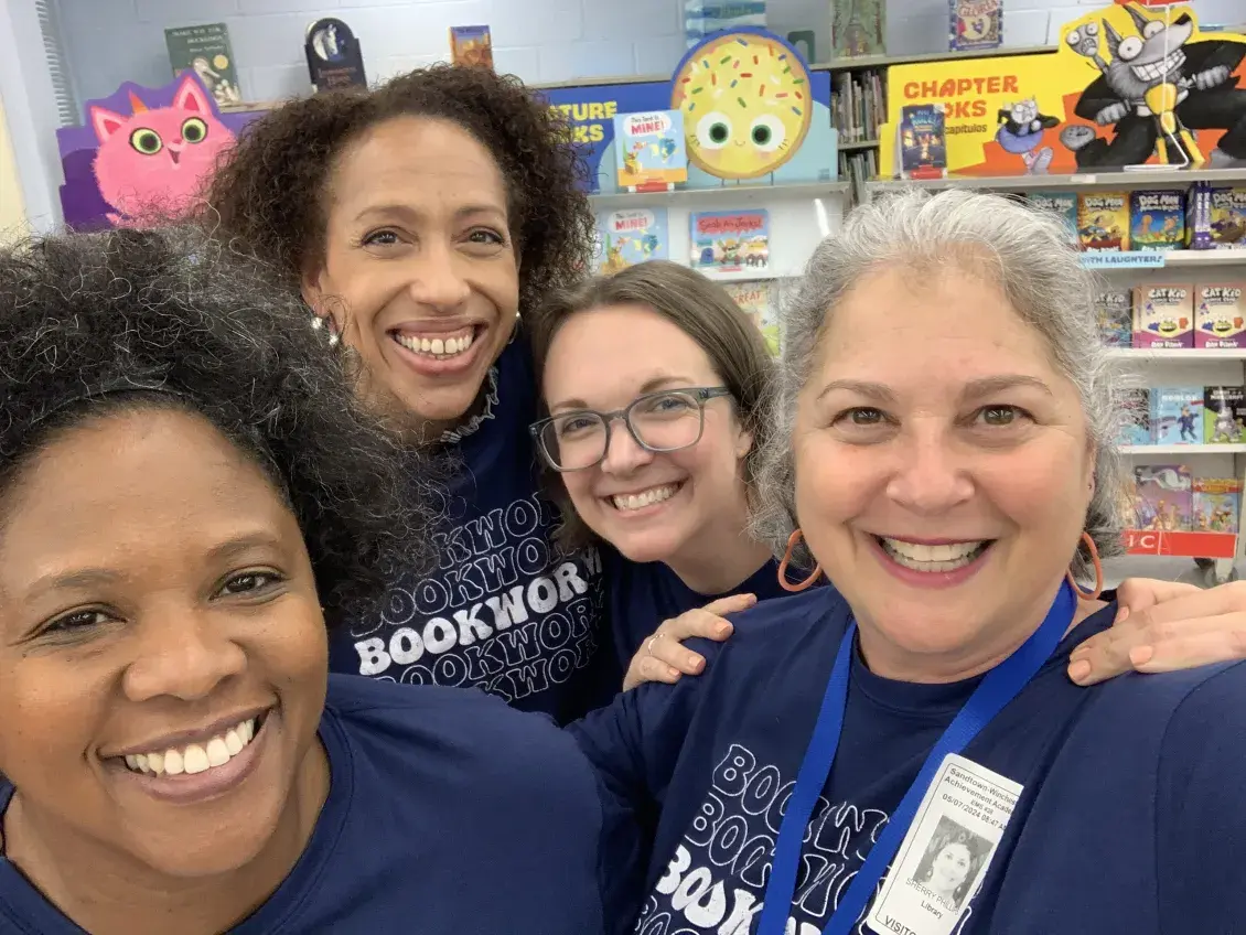 Group of four women stand next to each other smiling with children's bookcase in the background