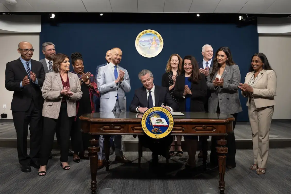 California Gov. Gavin Newsom sitting at desk signing a document, surrounded by clapping people