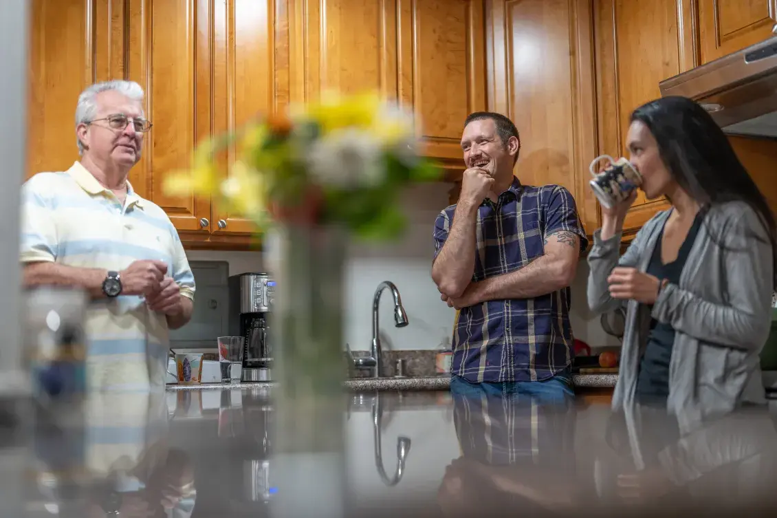 Three people stand in a kitchen smiling and talking with a vase with flowers in the center.
