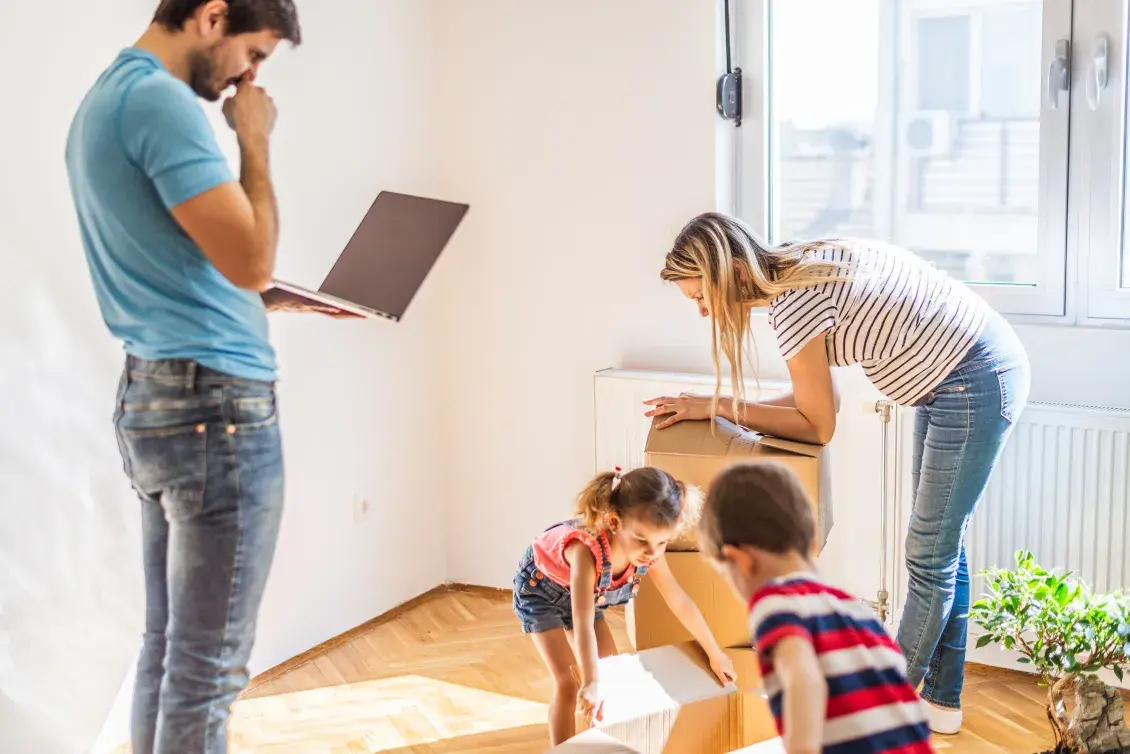 Family stands in sunlit room moving boxes while person stands staring at a laptop screen and looking uneasy