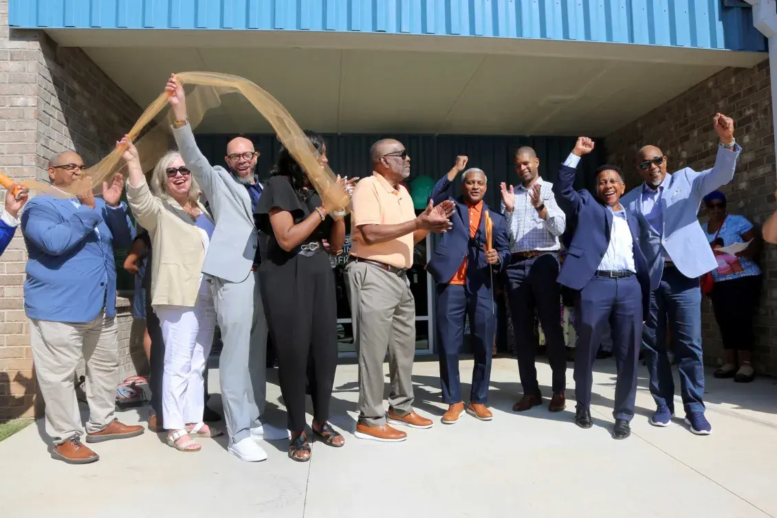 A group of people stand smiling and holding a ribbon in front of a building entrance