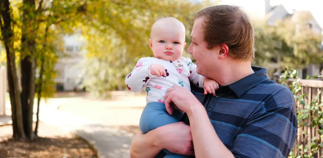 A father holds a baby on a sidewalk with trees and buildings in the background