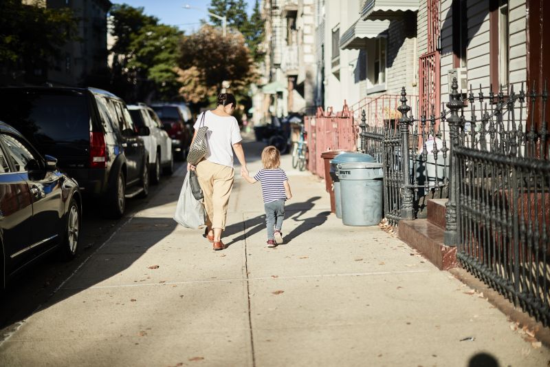 Mother and child walk on a neighborhood sidewalk along apartments, homes, and parked cars