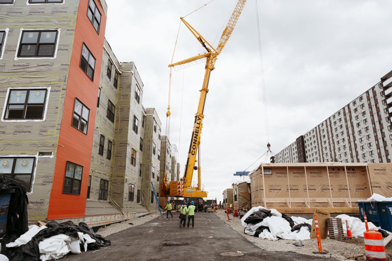 A crane sits between a modular housing project and one of its units, ready to be lifted and set in place.