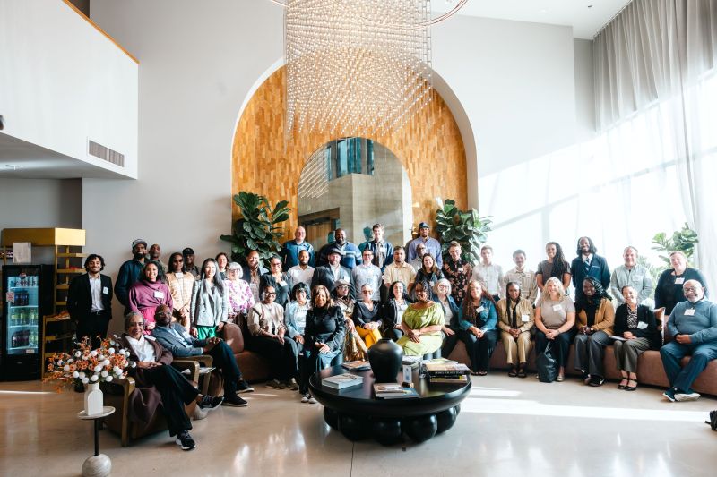A group of people pose together in a sunlit room with high ceiling in front of a white wall with a tan wooden arch panel