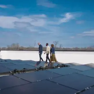 Three people walk alongside a rooftop solar array with a blue sky and clouds in the background