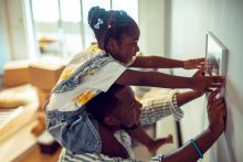 Girl sitting on her father's shoulders helps hang a picture in their home