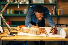 Man leans over wooden drafting table, working on an architectural model and designs