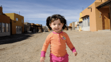 A little girl with an orange shirt smiling while standing on a construction site.