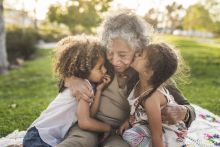 Older woman sits on blanket on the lawn with two small children. 