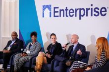 Five people sitting in chairs on stage having a discussion. The Enterprise logo is on the wall behind them. A man holds a microphone.