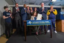 New York Governor seated at table holding legislation, surrounded by clapping group