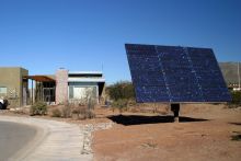 Desert setting with large ground-mounted solar panel in the forefront and a low-rise adobe-style apartment community in the background.