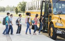 A line of children and youth carrying backpacks and brown lunch bags boards a yellow school bus.