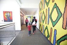 A mother and child with their backs to the camera walk down a hallway. On the right is a brightly colored wall painted with geometric shapes.