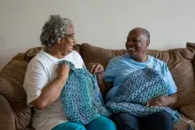 Two smiling women on couch holding up their colorful, blue crochet squares.