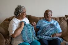 Two smiling women on couch holding up their colorful, blue crochet squares.