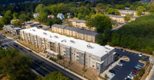 Arial view of 4-story multifamily apartment development with more housing and large trees in the background and a parking area to the left of the development.