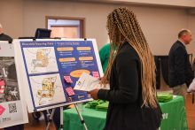 A young black woman is looking at a posterboard titled Riverdale Housing Data, with papers in her hand that have the Purple Line Corridor Coalition logo on them