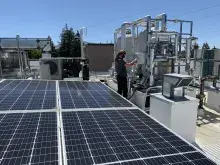 Rooftop view of flat solar panels with a worker adjusting a heat pump; blue sky and trees are in the background