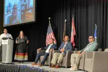 Men sitting in chairs with a woman and man standing behind a podium and a woman on a screen