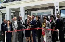 Smiling people in business attire standing in front of a building and cutting a red ribbon.