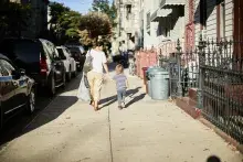 Mother and child walk on a neighborhood sidewalk along apartments, homes, and parked cars