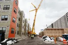 A crane sits between a modular housing project and one of its units, ready to be lifted and set in place.