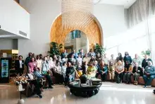 A group of people pose together in a sunlit room with high ceiling in front of a white wall with a tan wooden arch panel