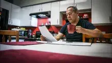 Man in dark shirt sitting at kitchen table drinking coffee and looking at paperwork