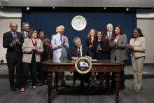 California Gov. Gavin Newsom sitting at desk signing a document, surrounded by clapping people
