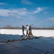 Three people walk alongside a rooftop solar array with a blue sky and clouds in the background