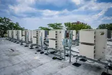 Row of rooftop heat pumps with trees, puffy white clouds, and blue sky in the background.