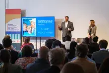 Two presenters address attendees from a stage while standing beside a presentation screen.