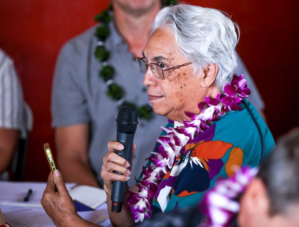 An older person speaks into a microphone with a phone in hand