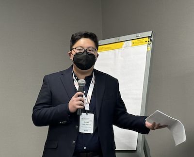 Man in black blazer and face mask with microphone speaking to the room 