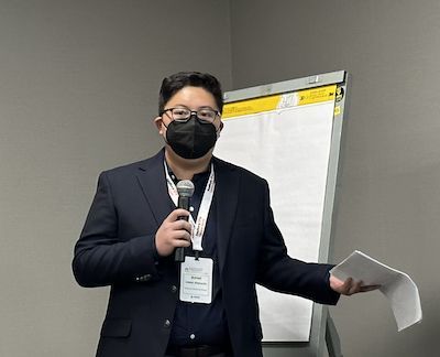 Man in black blazer and face mask with microphone speaking to the room