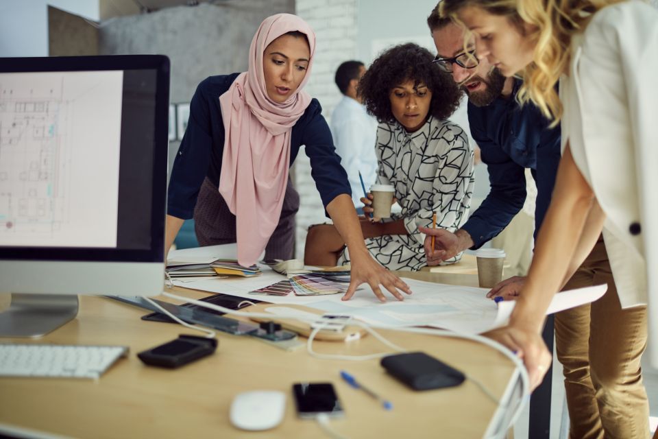 Woman in a pink head scarf stands at a table reviewing documents with a group of people. A computer monitor sits on the table on the woman's left.