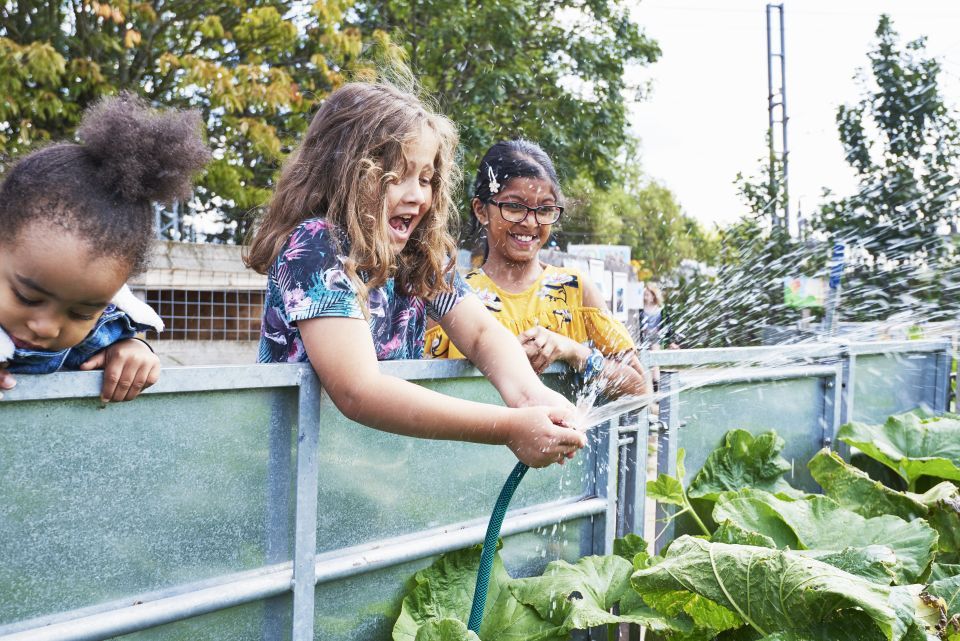 A young girl in a blue top delights in watering a garden with a hose. Two more young girls stand to her right and left watching..