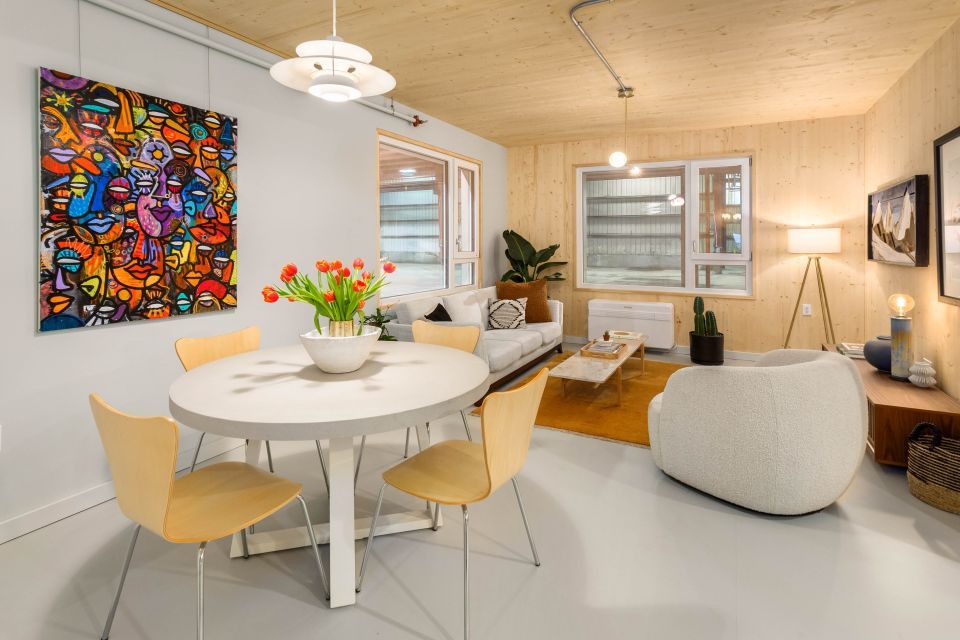 Inviting interior of a home featuring natural wood grains and a colorful painting on the wall to the left. A vase with orange flowers is on a round dining table.