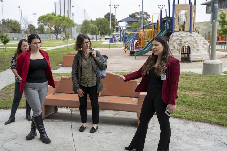 Woman with long dark hair wearing a red blazer speaks to a small group of people. A playground is in the background.
