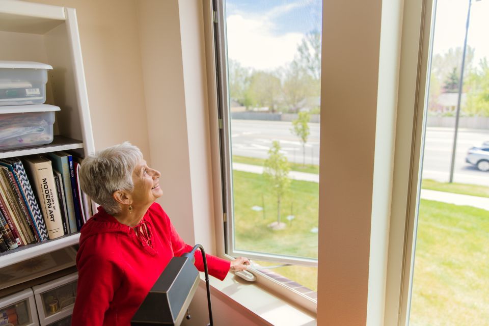 Woman with short silver hair wearing a red shirt stares up as she uses a crank handle to open the casement window.