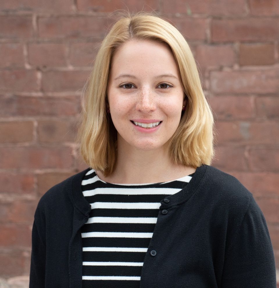 Person with shoulder-length blond hair wearing a black-and-white striped top and a black cardigan, with a brick wall in the background.