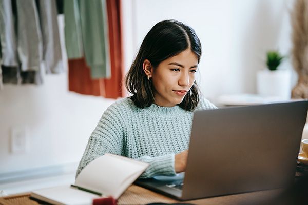 A woman works at a laptop with a notebook and pen on the desk