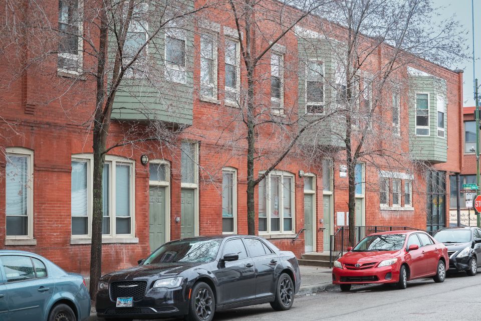 A row of two-story brick townhomes with sage-colored doors and parked cars on the street level.