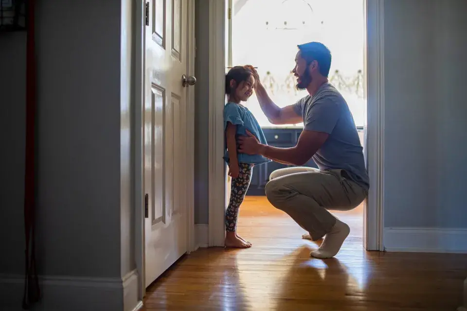 Young girl stands against the wall as her father squats in front of her to measure her height