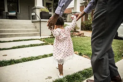 Toddler walks on a sidewalk assisted by a parent