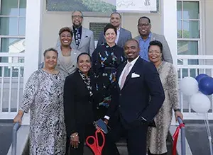 A group of people stand on the stairs of a home, and a woman holds a large pair of scissors